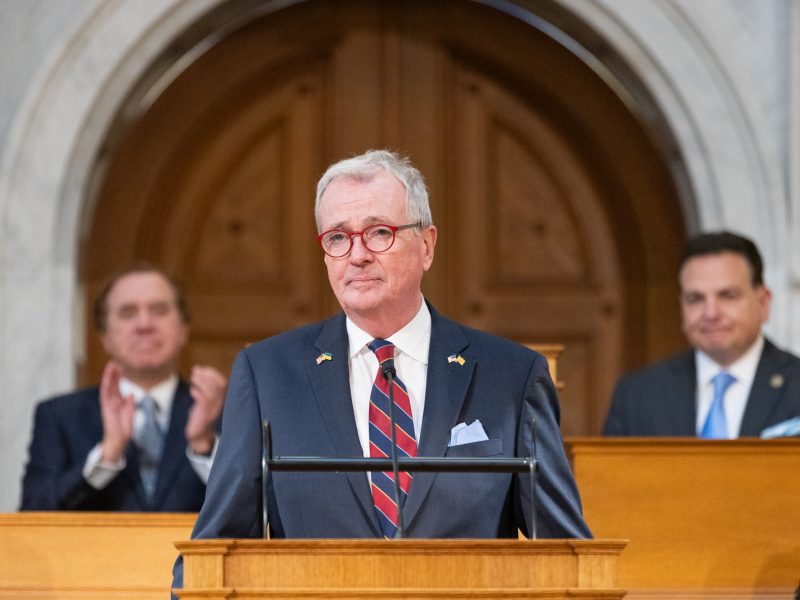 Gov murphy stands at a podium as craig coughlin and nick scutari sit behind him
