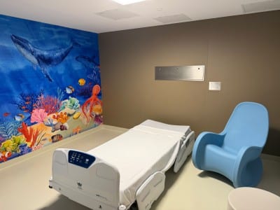 Hospital room with wall that shows a picture of an underwater scene. in the foreground there's a hospital bed and blue chair.
