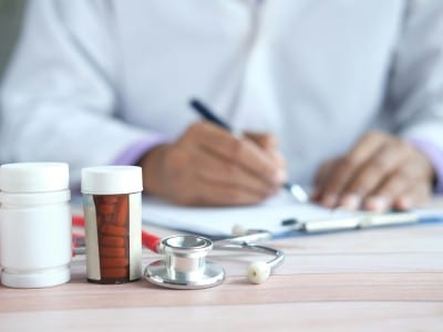 Person in white lab coat writes something. A pair of glasses and prescription medication is in front of them.