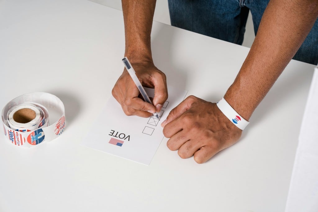 Person's hands on table. They're holding a pen and marking off a piece of paper that says VOTE