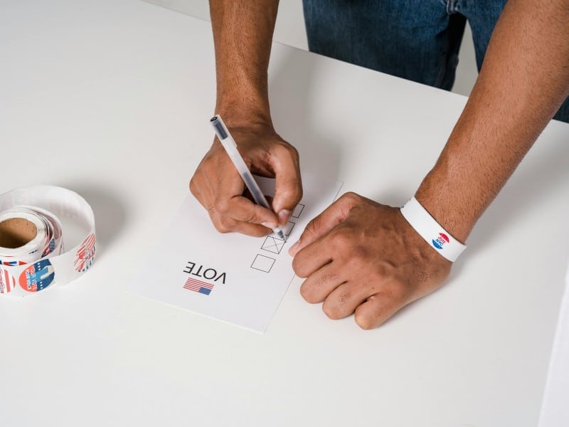 Person's hands on table. They're holding a pen and marking off a piece of paper that says VOTE
