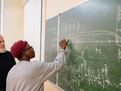 A student writes complex mathematical equations on a chalkboard during a university lecture.